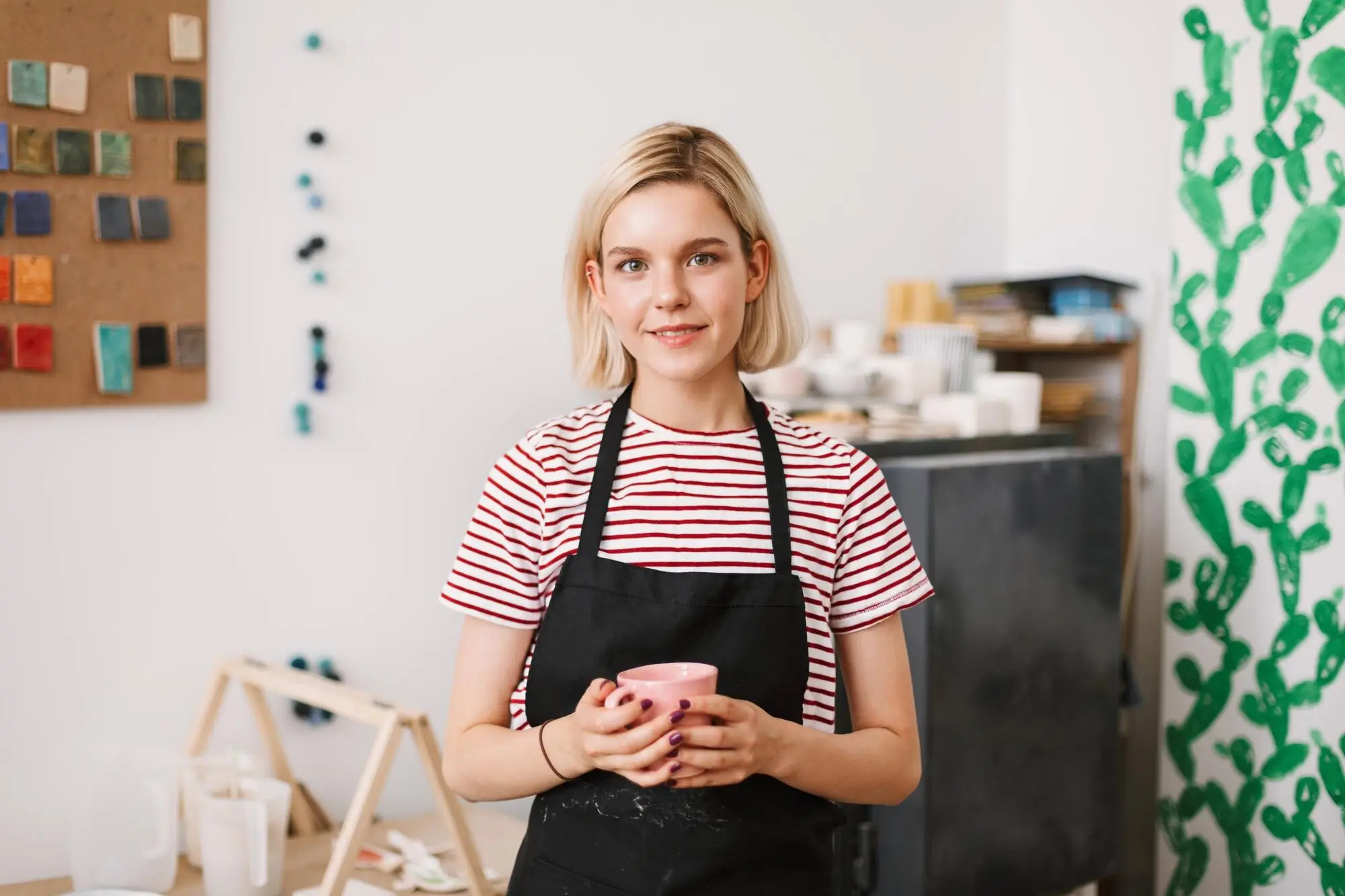 In einer Töpferwerkstatt hält ein hübsches Mädchen in schwarzer Schürze eine handgefertigte Tasse in den Händen und blickt verträumt in die Kamera, praxisnah und nachvollziehbar.