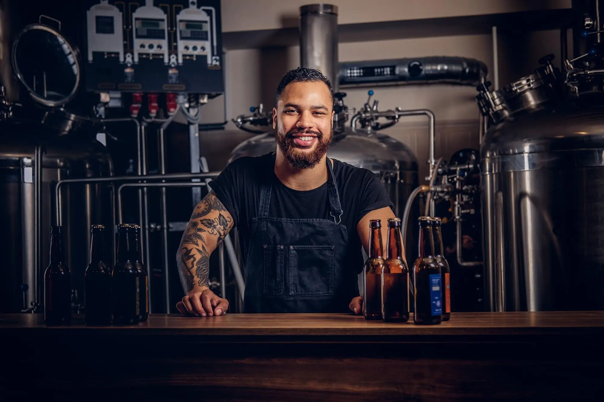 Porträt eines stilvollen, bärtigen, dunkelhäutigen Mannes mit einem Tattoo auf der Hand, der hinter der Theke in einer Brauerei steht, praxisnah und nachvollziehbar.